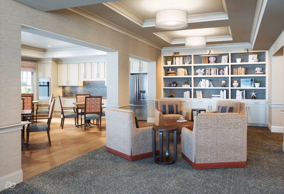 A cozy common area in a senior living facility featuring four upholstered armchairs arranged around a small round wooden table on a carpeted floor. Behind the seating area is a built-in bookshelf filled with books and decorative items. To the left, there is an open view into a dining area with tables and chairs, and a kitchen with white cabinetry and stainless steel appliances.