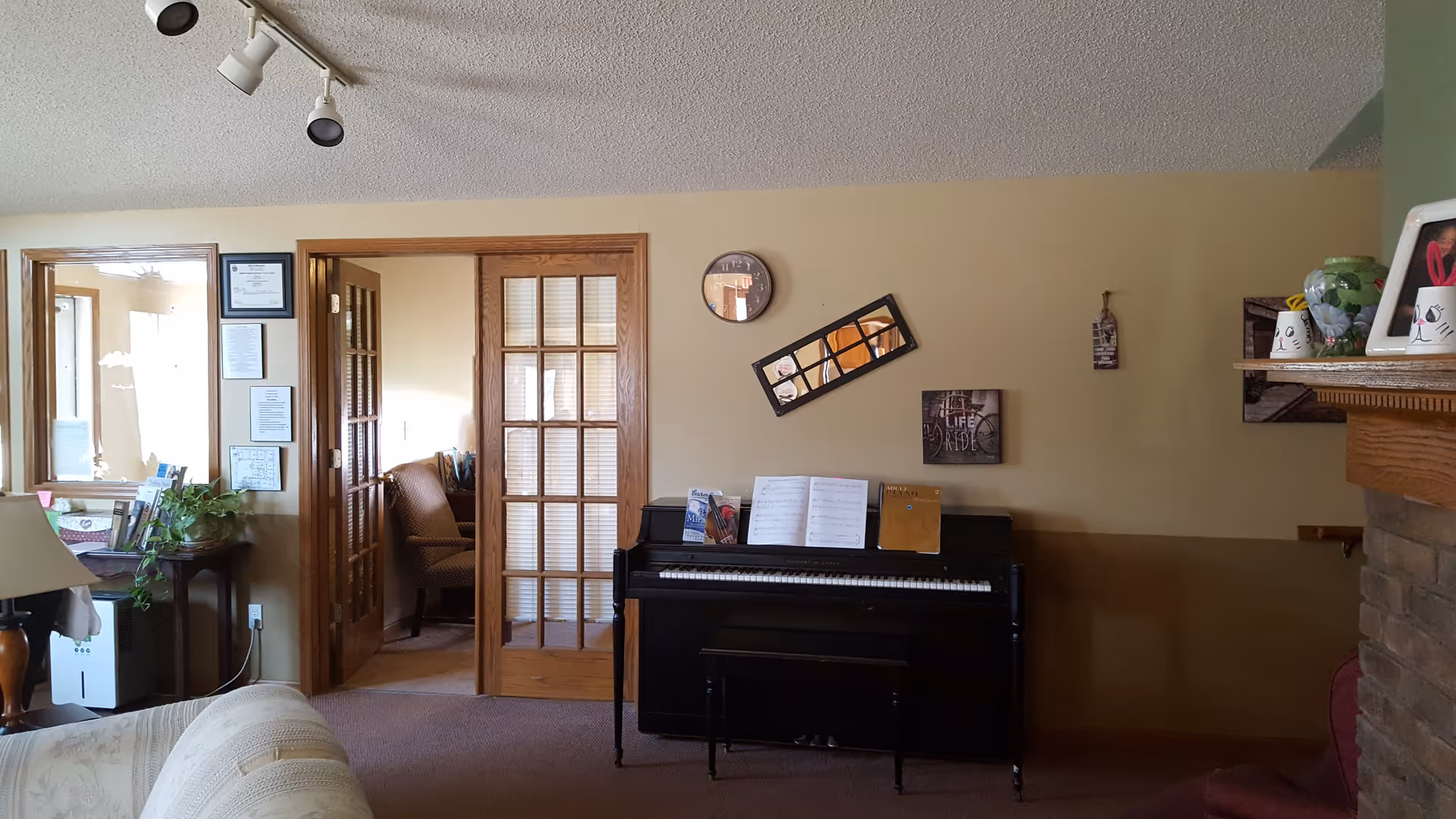 Interior view of a living room area with a black upright piano against a beige wall. Above the piano, there is a round clock, a tilted rectangular mirror, and a small framed picture. To the left, there are wooden French doors leading to another room with a patterned armchair. A small table with a green plant and some papers is near the doors. The right side shows part of a brick fireplace mantel with decorative items on top. The ceiling has track lighting with three spotlights.