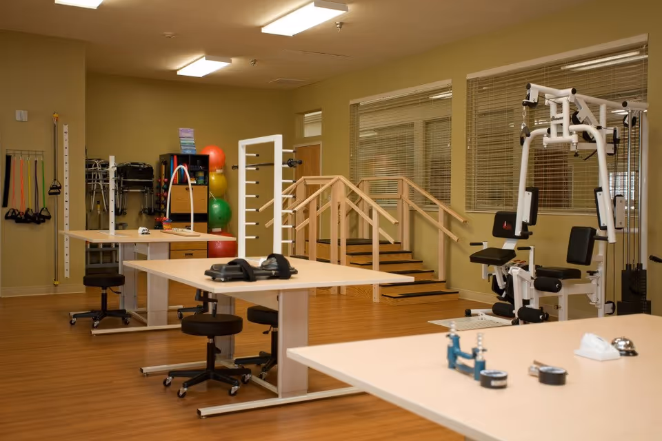 A physical therapy room with exercise equipment including resistance bands, therapy tables with stools, a set of stairs with handrails for rehabilitation, colorful exercise balls, and a multi-function weight machine. The room has wooden flooring and beige walls with windows featuring blinds.