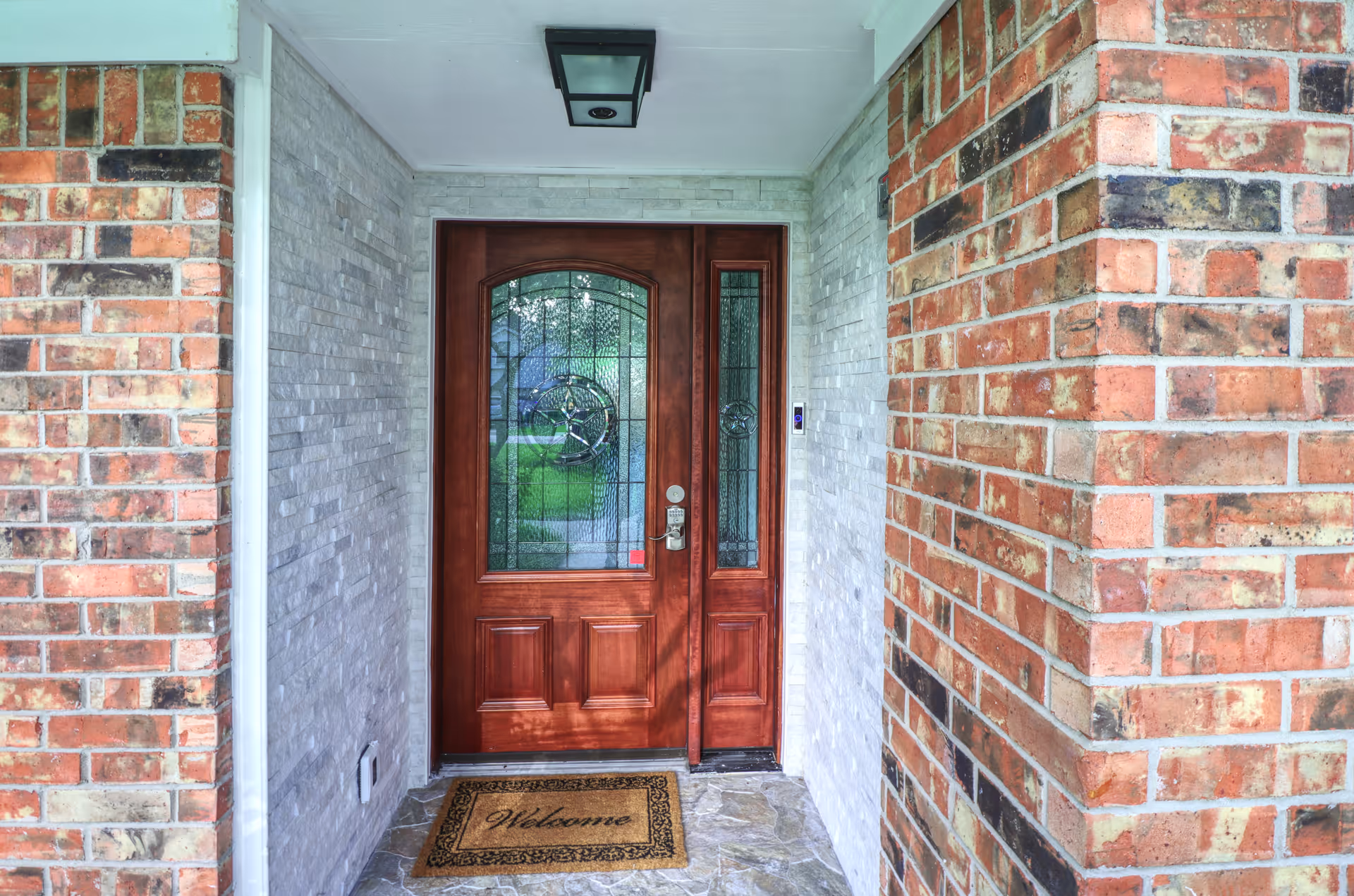 Front entrance of a building with a wooden door featuring decorative glass panels. The entrance is framed by brick walls on either side and light-colored stone walls behind. A welcome mat is placed on the stone floor in front of the door, and a ceiling light fixture is mounted above the door.