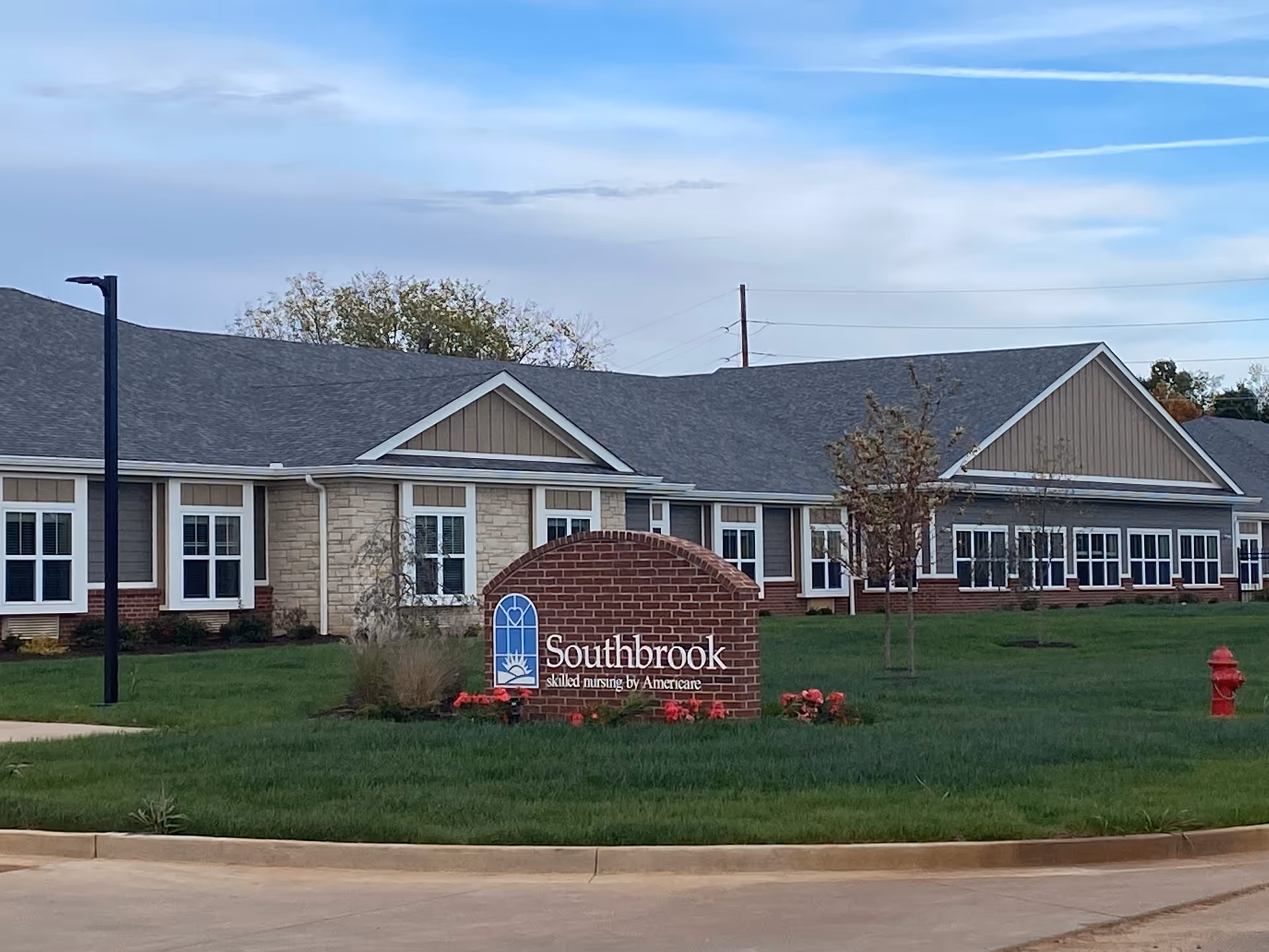 Exterior view of a single-story building with multiple windows and a gray roof. In front of the building, there is a brick sign with the text 'Southbrook skilled nursing by Americare' surrounded by green grass and some small plants and flowers. The sky is partly cloudy.