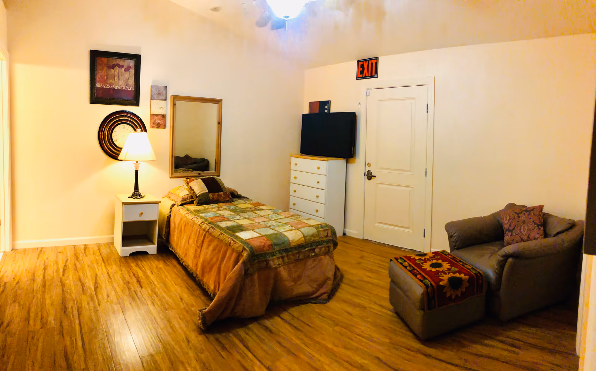 A cozy bedroom with a single bed covered in a patchwork quilt, a nightstand with a lamp, a wall clock, framed artwork, a mirror, a dresser with a TV on top, and a comfortable armchair with an ottoman. The room has wooden flooring and an exit sign above the door.