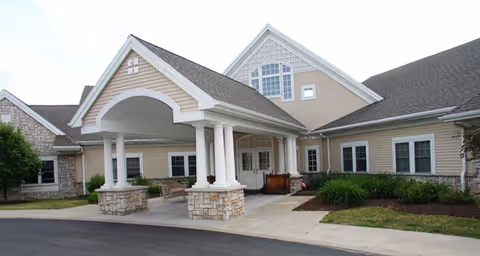 Exterior view of a single-story senior living facility building with beige siding and stone accents. The entrance features a covered driveway supported by white columns, with a paved driveway and landscaped greenery around the building.
