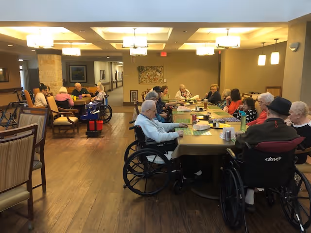 A group of elderly people, some in wheelchairs, sitting around two large tables in a well-lit common area of a senior living facility. The room has wooden floors, beige walls, and modern ceiling lights. Some individuals are engaged in conversation or activities.