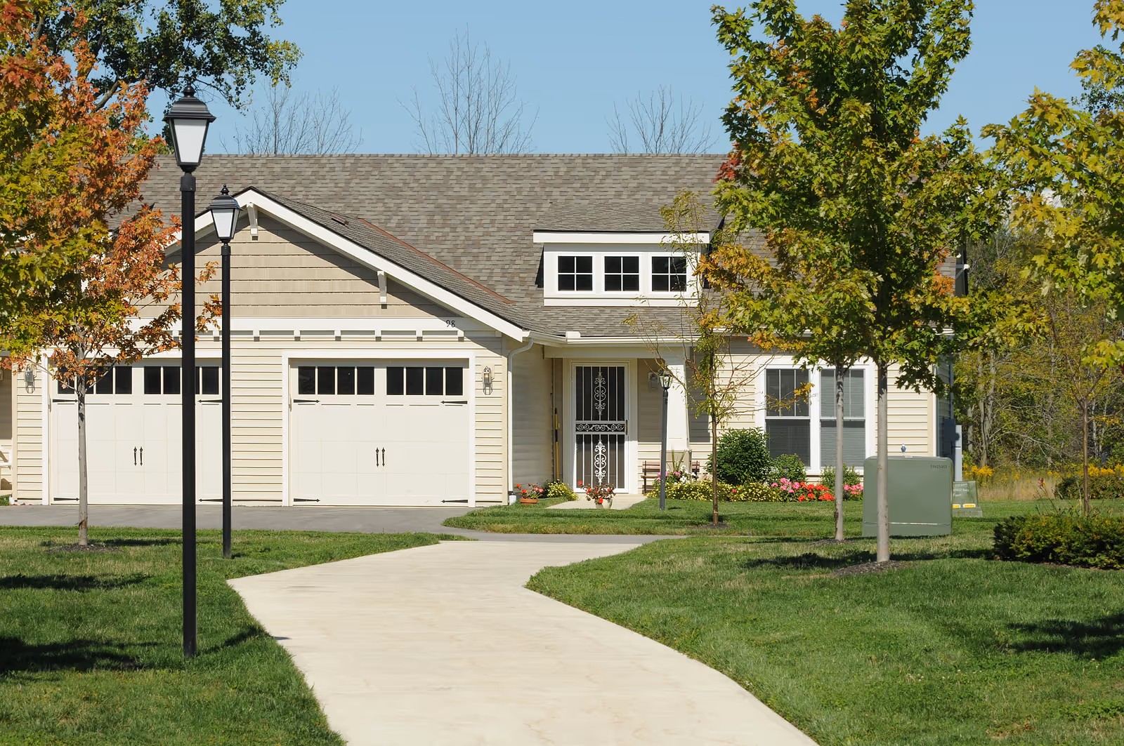 Front view of a single-story beige house with a double garage, walkway, lamp posts, and landscaped lawn.