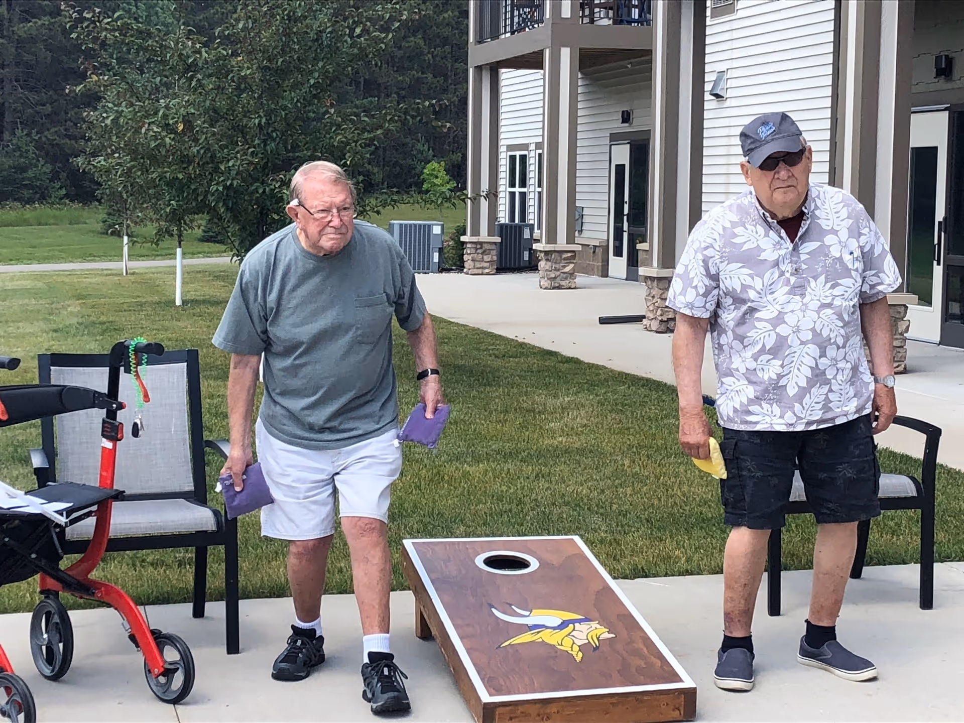 Two elderly men playing cornhole outside on a concrete patio next to a building. One man is wearing a gray t-shirt and white shorts, holding purple bean bags, and the other man is wearing a floral shirt, black shorts, and a cap, holding a yellow bean bag. There are chairs and a red walker nearby, with grass and trees in the background.