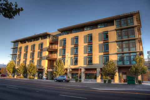 Front exterior of a multi-story senior living building with trees and parked cars along the street.