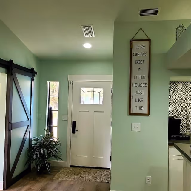 Light-green painted foyer with a white front door, a dark barn-style sliding door, a potted plant, and a hanging wall sign next to a view into the kitchen.