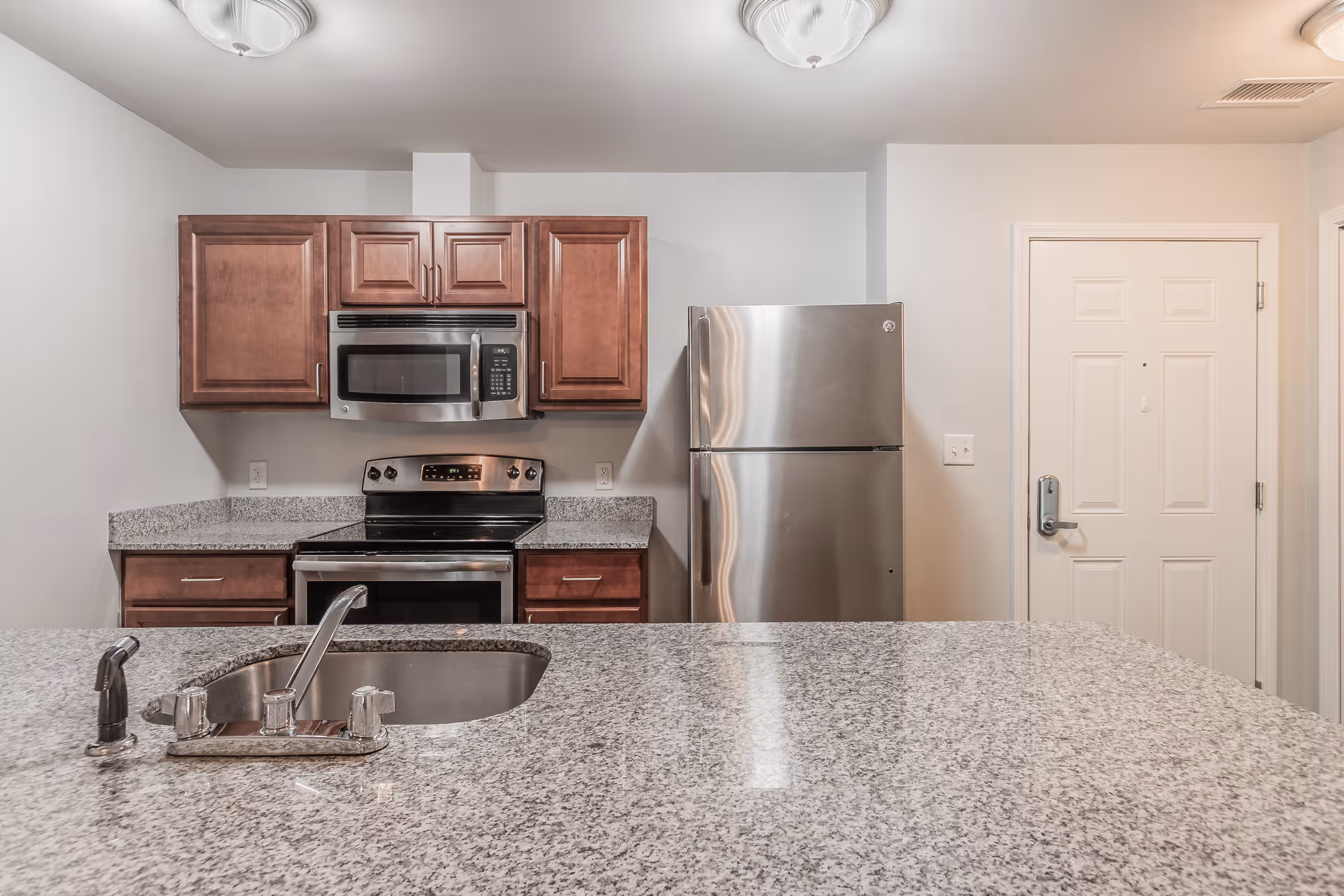 Modern kitchen with granite countertops, a stainless steel sink with faucet, wooden cabinets, a stainless steel microwave above a stove, and a stainless steel refrigerator next to a white door.