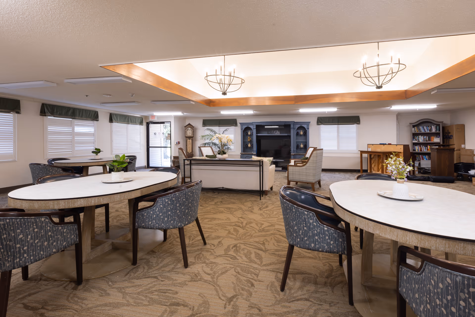 Spacious communal dining and activity room with round tables and chairs, a seating area and TV cabinet at the far end.