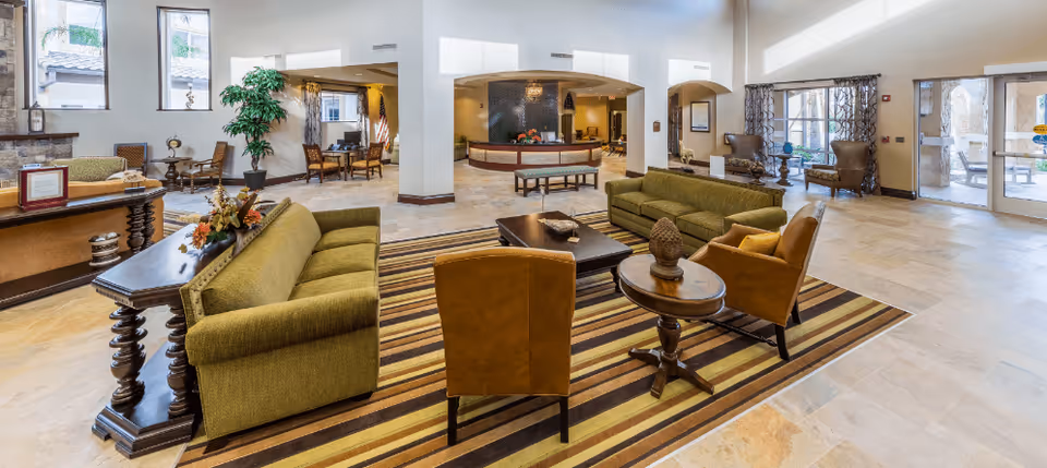 Spacious senior living facility lobby with green sofas, brown armchairs, a striped area rug, and a reception desk in the background. Large windows and glass doors allow natural light to fill the room, and there are decorative plants and framed artwork on the walls.