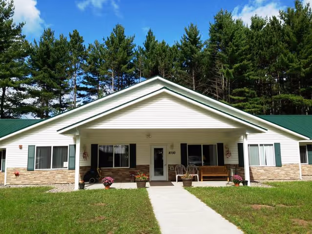 Front exterior view of a single-story building with white siding, green roof, and a covered entrance. There are potted plants and outdoor seating on the porch, surrounded by green grass and tall pine trees in the background under a blue sky with some clouds.