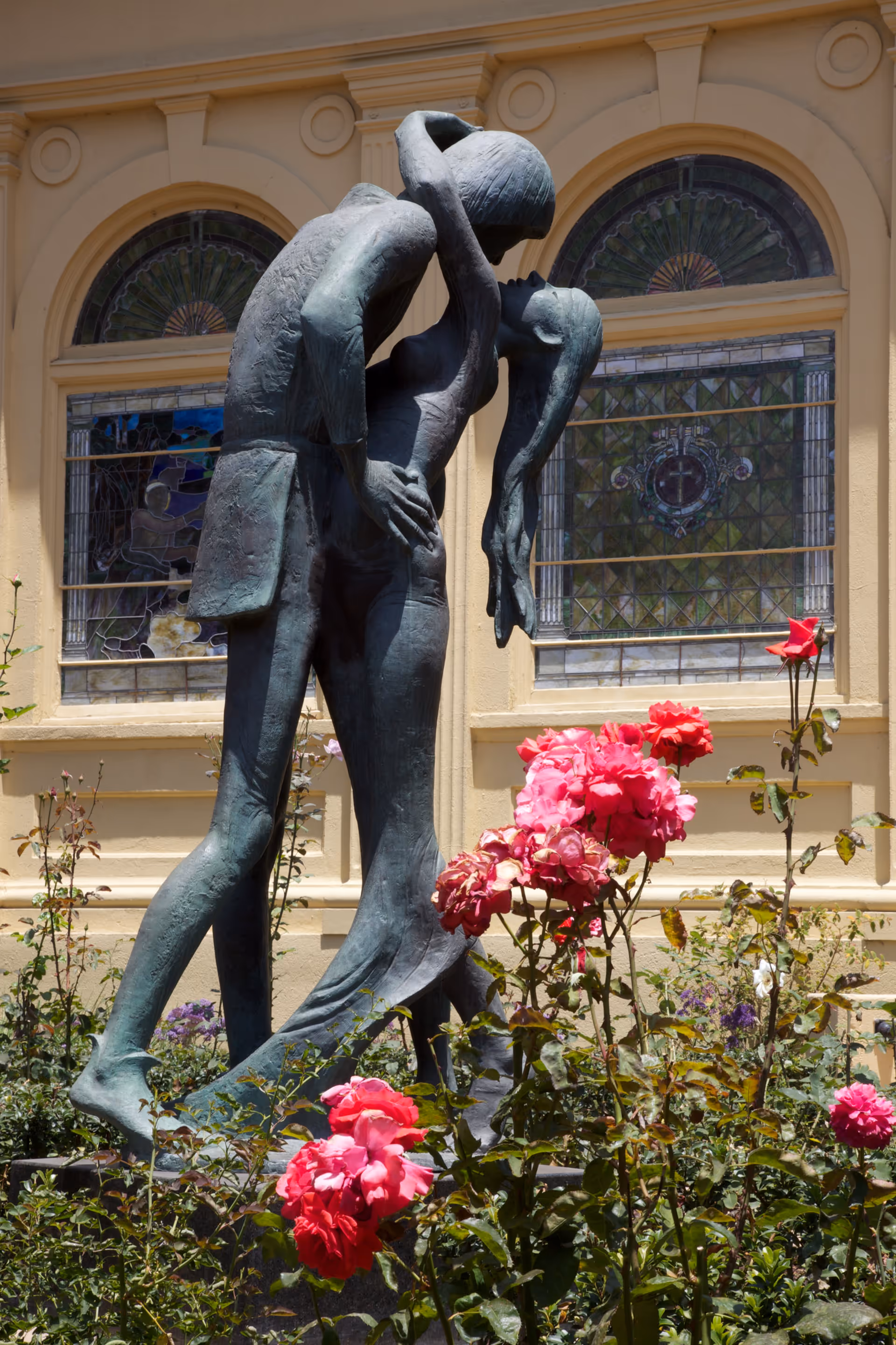Bronze sculpture of a couple embracing and kissing, surrounded by blooming pink roses, with a building featuring stained glass windows in the background.