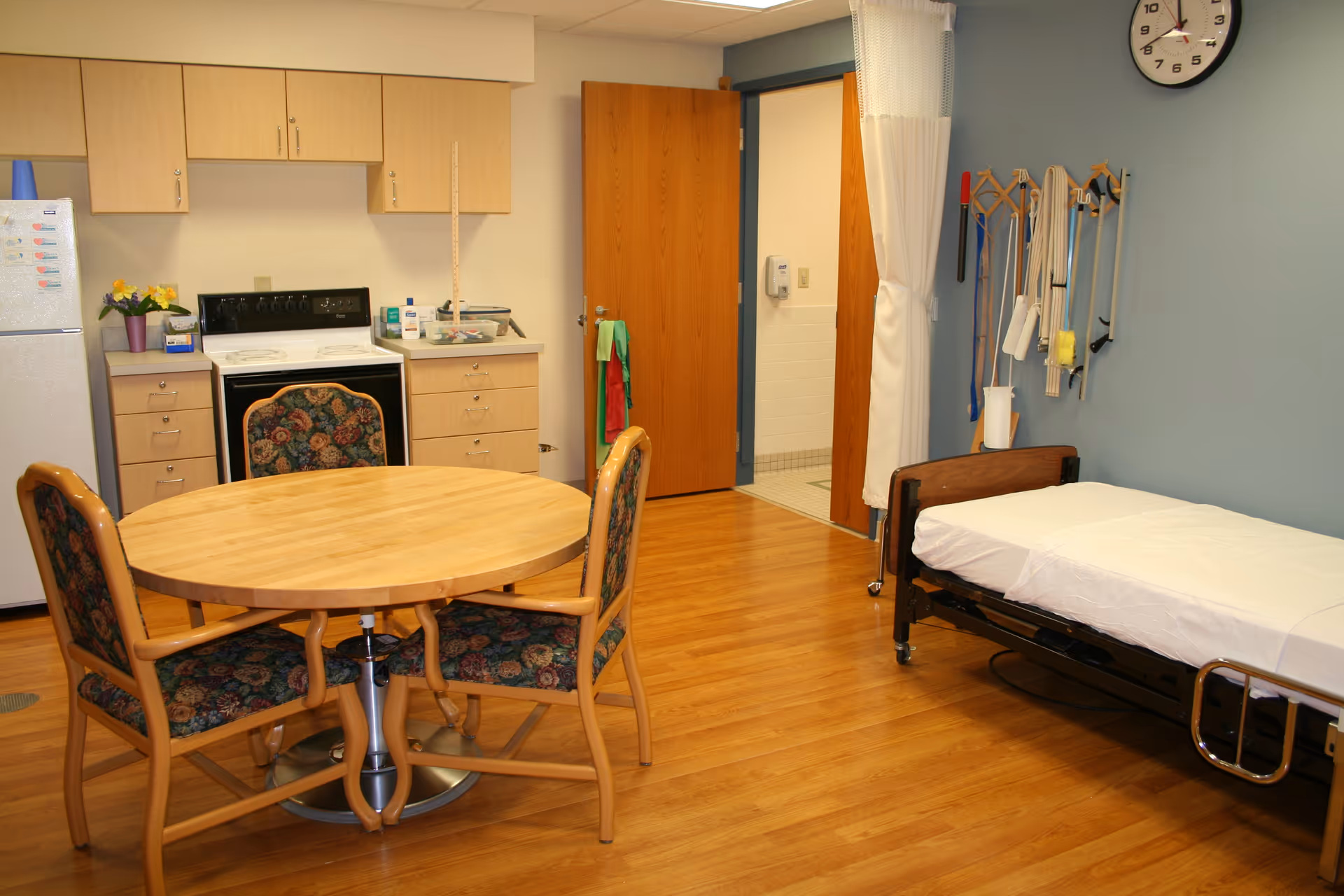 A room with a small kitchen area including a refrigerator, stove, and cabinets. In the center is a round wooden table with four floral upholstered chairs. To the right is a hospital-style bed with white sheets and a blue wall behind it. Various medical or therapy equipment hangs on the wall near the bed. A door is open leading to a tiled bathroom area.