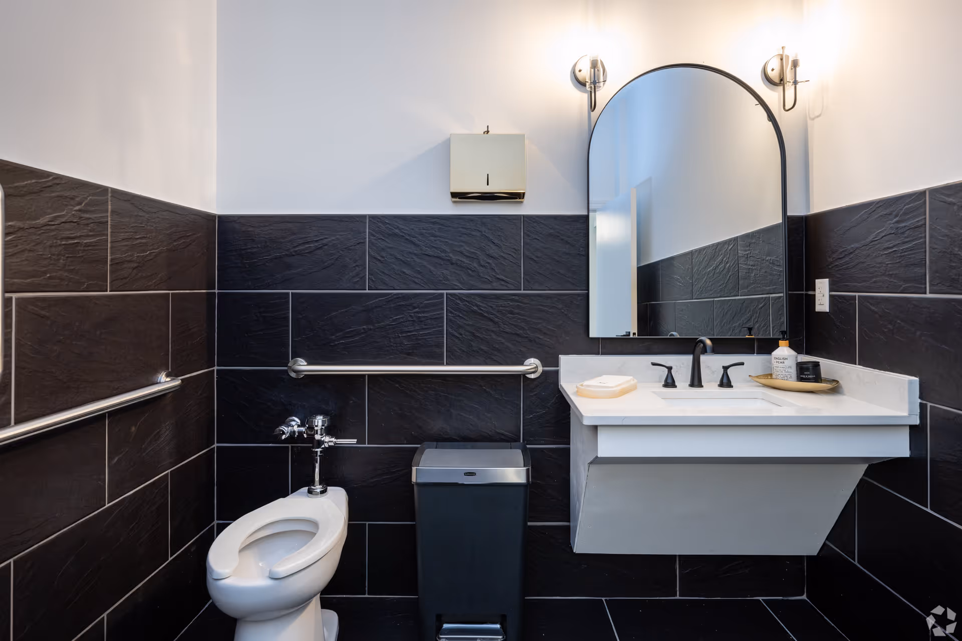 A modern bathroom with black textured tiles on the walls and floor. The bathroom features a white toilet with a safety grab bar on the wall beside it, a black trash bin, and a white floating sink with black faucets. Above the sink is a large arched mirror flanked by two wall-mounted lights. A soap dispenser and a small tray with toiletries are placed on the sink countertop.