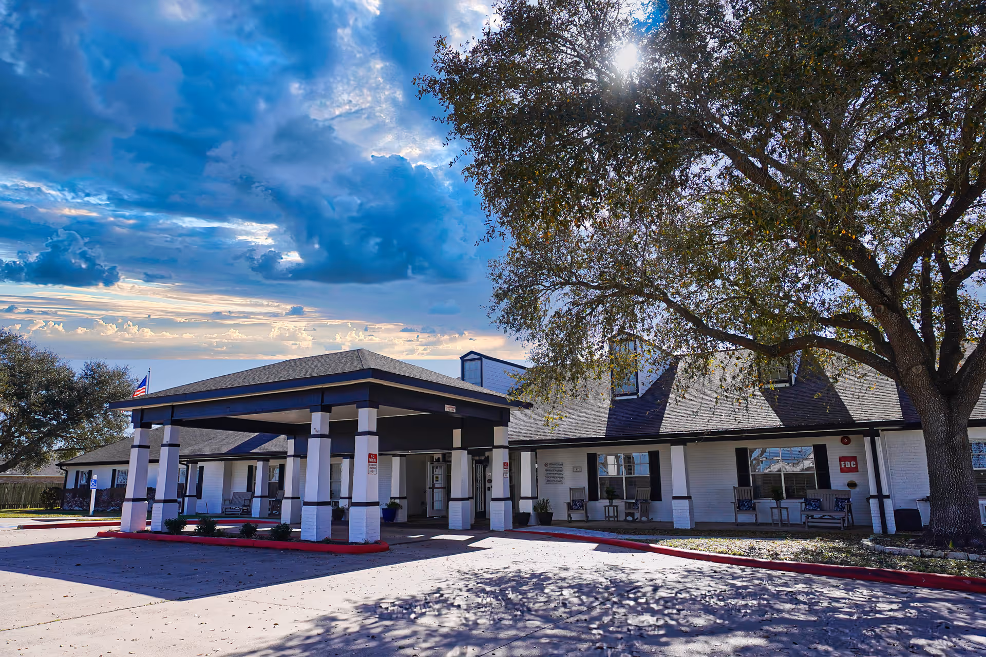 Exterior view of Sodalis Victoria Assisted Living facility showing a single-story building with a covered entrance supported by white columns. There is a large tree casting shadows on the driveway and a partly cloudy sky with the sun shining through.