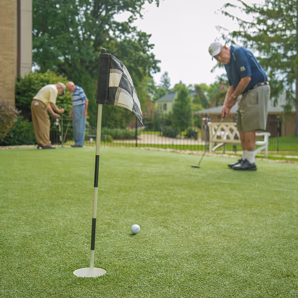 Three elderly men playing golf on a putting green outdoors, with one man putting near the hole marked by a black and white checkered flag, and two others standing and preparing in the background surrounded by trees and residential buildings.