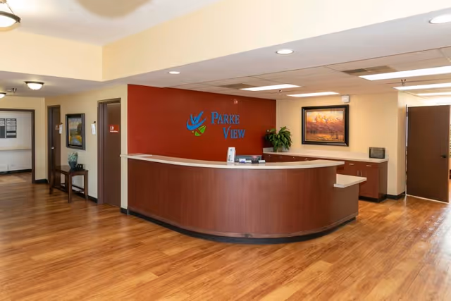Reception area of Parke View Rehabilitation & Care Center with a curved wooden front desk, a red accent wall behind the desk featuring the facility's logo, wooden flooring, a framed painting on the wall, and doors leading to other rooms.