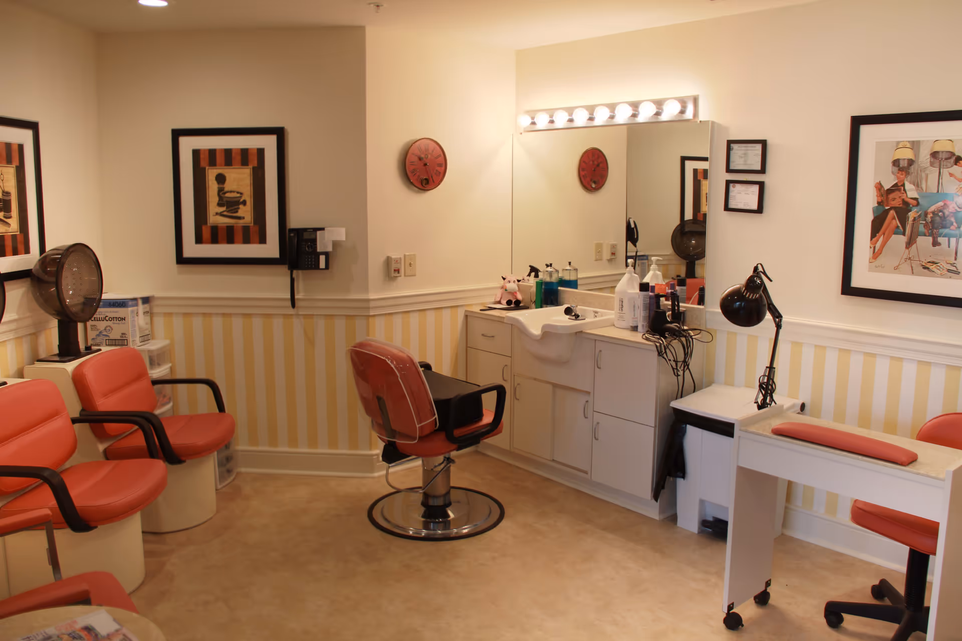 A small salon area in a senior living facility with orange salon chairs, a large mirror with lights above, a sink, various hair care products, and framed artwork on the walls. There is also a manicure station with a lamp and a padded armrest.