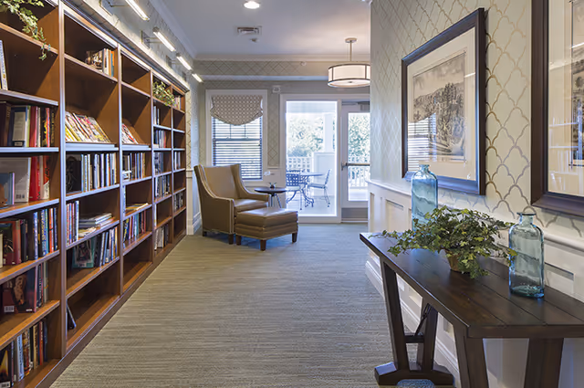 A bright interior reading area with bookshelves along the left wall, a leather armchair and small table near a glass door to a patio, and a console table with framed art and vases on the right.