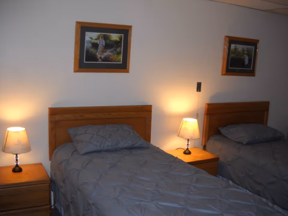 Two twin beds with gray bedding and wooden headboards, flanked by nightstands with lamps and framed pictures on a beige wall.
