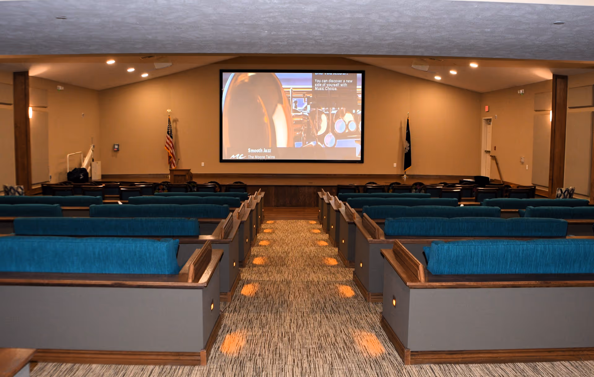 Interior view of a community room or auditorium with rows of teal cushioned pew-style seating facing a large projection screen. The room has beige walls, carpeted floor with lights along the aisle, and flags on either side of the screen.