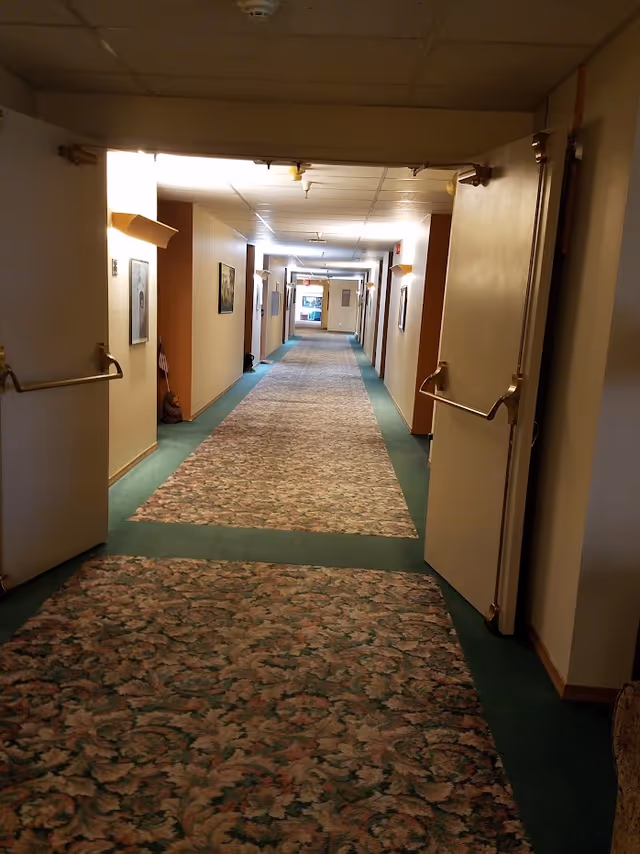 Long indoor hallway in a retirement community with patterned carpet and green borders, beige walls with framed pictures, and open double doors with brass push bars.