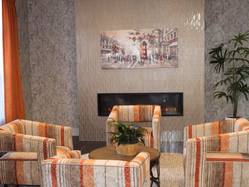 A cozy seating area with four striped upholstered armchairs arranged around a round wooden coffee table with a potted plant. Behind the chairs is a modern rectangular fireplace set into a textured wall with a painting above it depicting a street scene with a red telephone booth. There is a tall green plant in a white pot to the right and an orange curtain to the left.