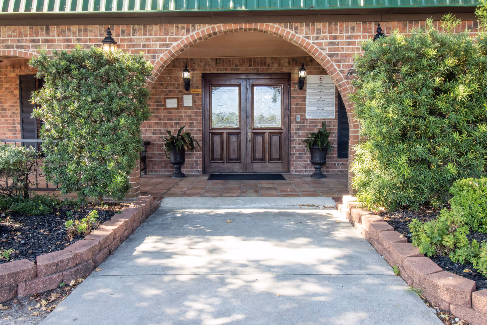 Entrance to a building with double wooden doors under a brick archway, flanked by two large green bushes and two black urn planters with plants. The building has a brick exterior and a green roof edge, with two wall-mounted lantern lights on either side of the doors.