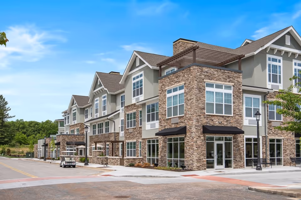 Exterior view of a modern senior living facility building with stone and siding facade, multiple large windows, and a clear blue sky above. The building has three stories and features outdoor seating areas and street lamps along the sidewalk.
