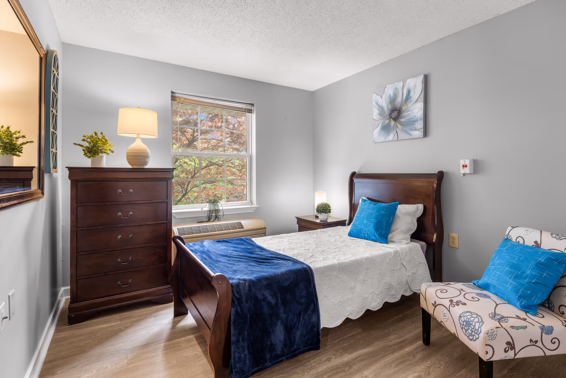 Sunlit private bedroom with a single wooden sleigh bed made up in white linens and blue accents, a dark wood dresser, patterned armchair, and a window overlooking trees.