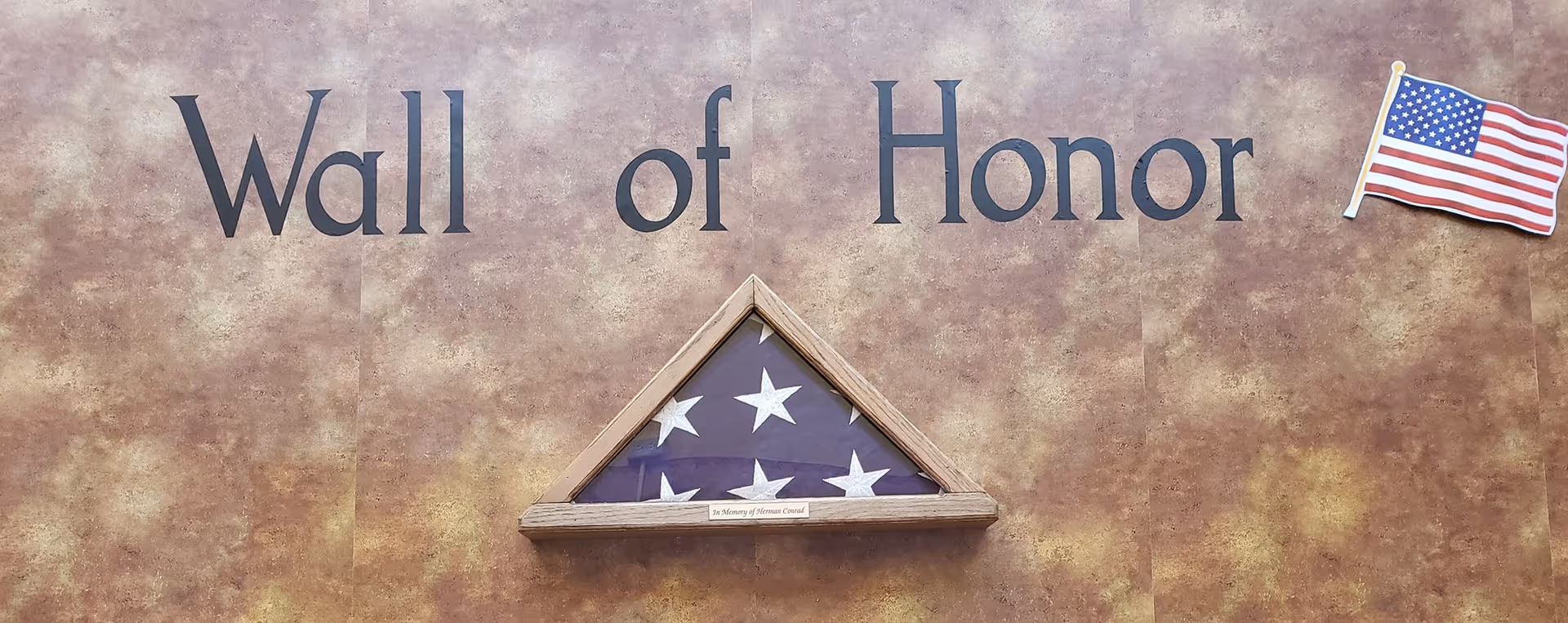 A wall display titled 'Wall of Honor' featuring a folded American flag in a triangular wooden case mounted on a textured brown background, with a small American flag graphic on the right side.