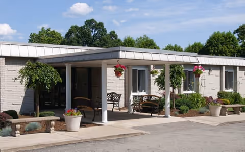 Single-story brick rehabilitation and healthcare center front with a covered entrance, benches, hanging flower baskets, and potted plants.