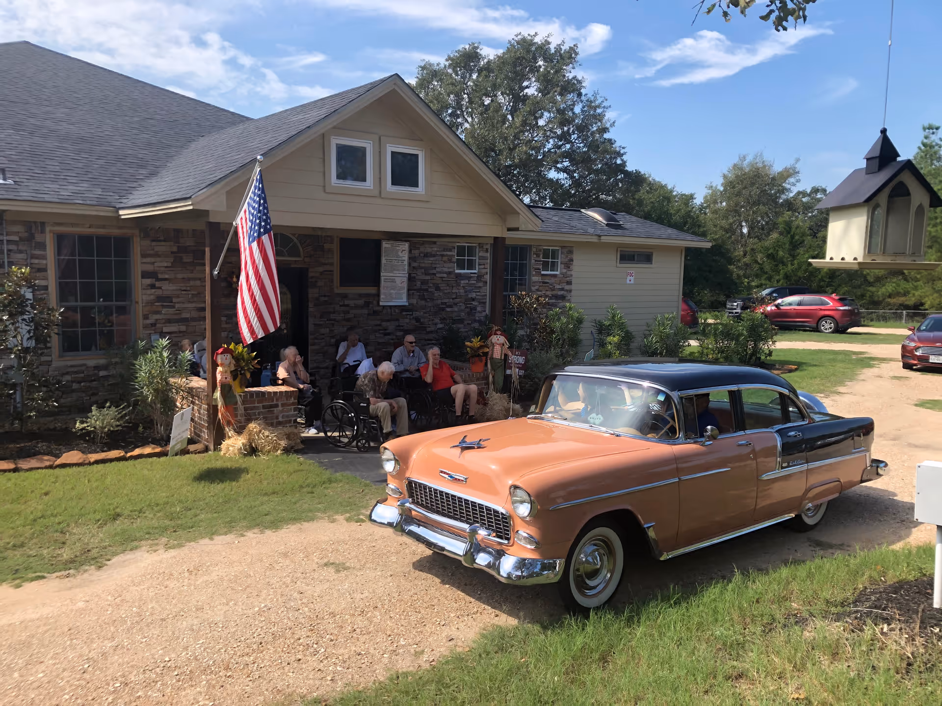 A group of elderly people sitting outside a brick and siding building with an American flag displayed on the porch. A vintage two-tone car is parked on a gravel driveway in front of the building. There is a birdhouse hanging on the right side of the image and several cars parked in the background under a partly cloudy sky.