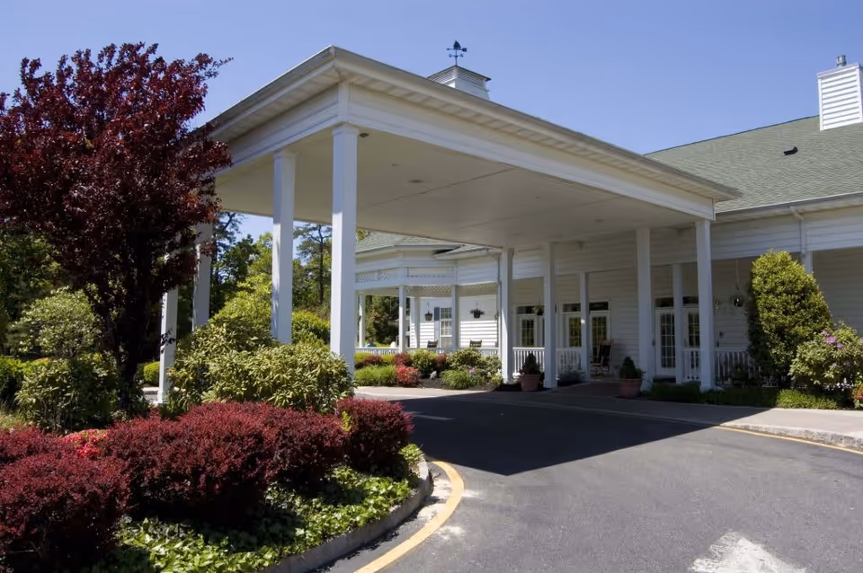 Entrance of a senior living facility with a covered drop-off area supported by white columns, surrounded by well-maintained landscaping including bushes and trees under a clear blue sky.