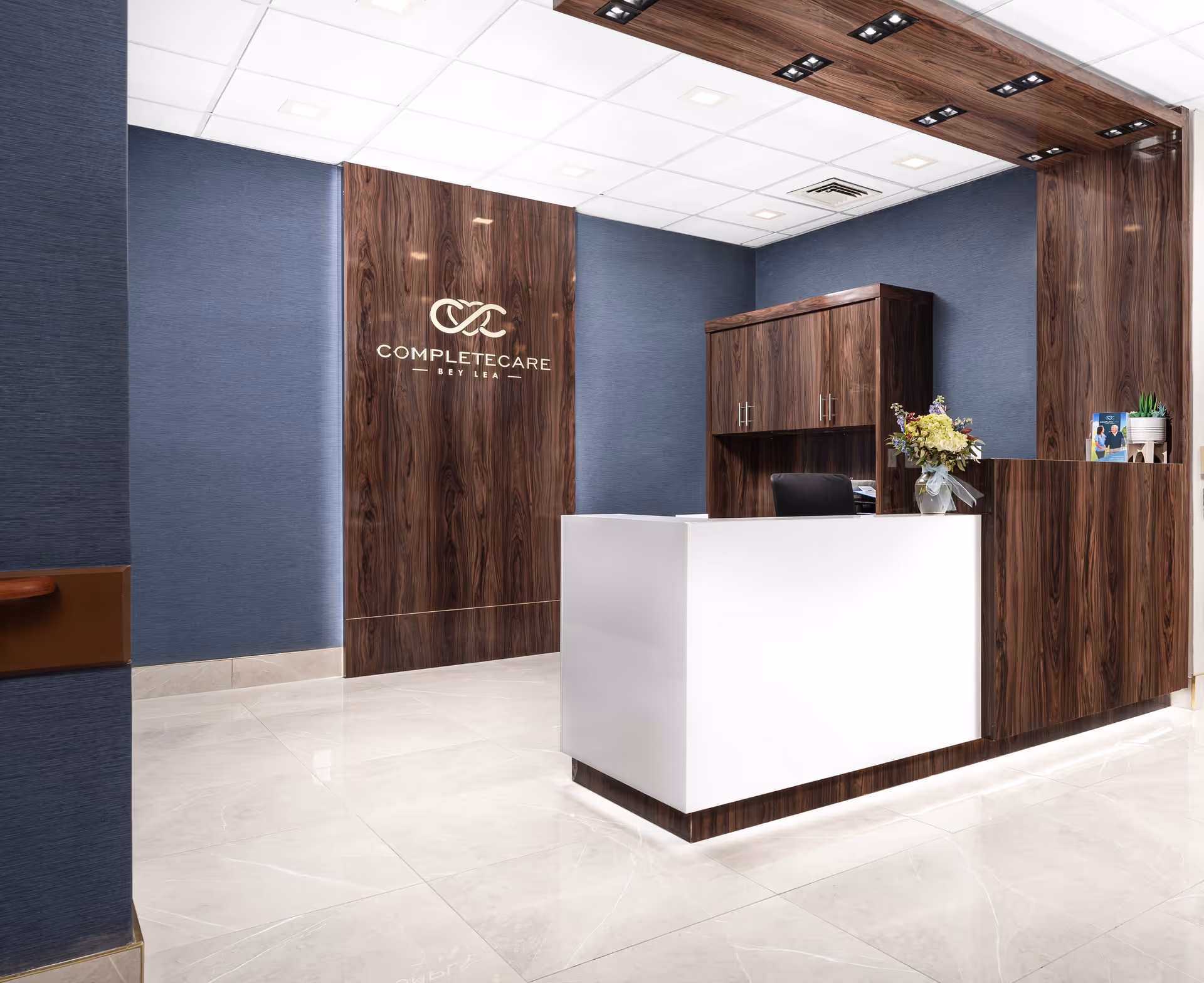 Reception area of Complete Care at Bey Lea featuring a modern white and wood-paneled front desk, a wood-paneled wall with the facility's logo, blue textured walls, a cabinet behind the desk, and a vase with flowers on the counter.