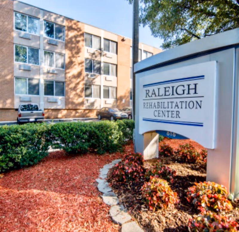 Outdoor view of Raleigh Rehabilitation Center showing a landscaped garden area with bushes and flowers in front of a multi-story brick building with several windows and parked cars.