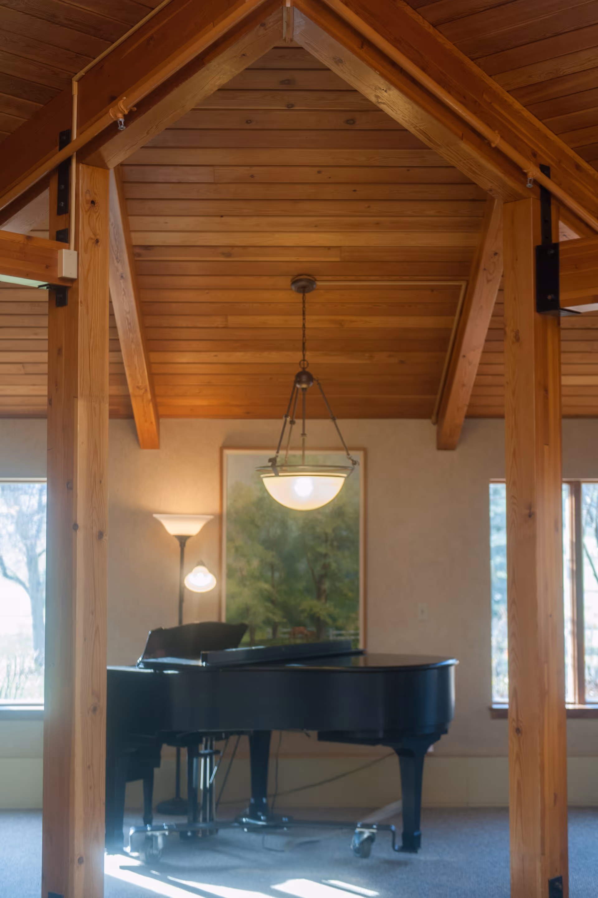 Interior view of a room with a wooden vaulted ceiling supported by wooden beams. A black grand piano is positioned in the center of the room, with a floor lamp and a hanging ceiling light above it. There is a framed picture of trees on the wall behind the piano and windows on either side letting in natural light.