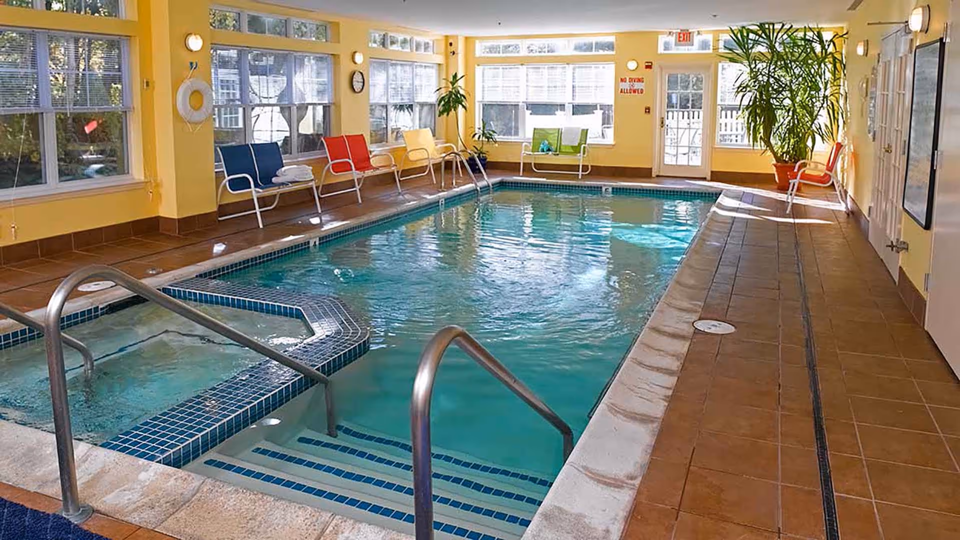 Indoor swimming pool area with a hot tub section, surrounded by yellow walls and large windows letting in natural light. Several colorful chairs are lined up along the walls, and there are potted plants near the windows and door. The floor is tiled and there are metal handrails leading into the pool.