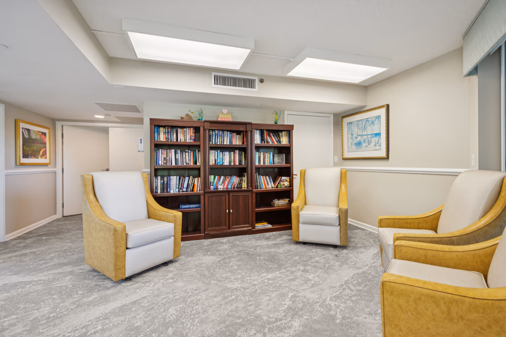 A cozy interior room with four beige and yellow armchairs arranged around a wooden bookshelf filled with books. The room has light-colored walls, a framed picture on the right wall, and a carpeted floor. The ceiling has two large rectangular fluorescent light fixtures.
