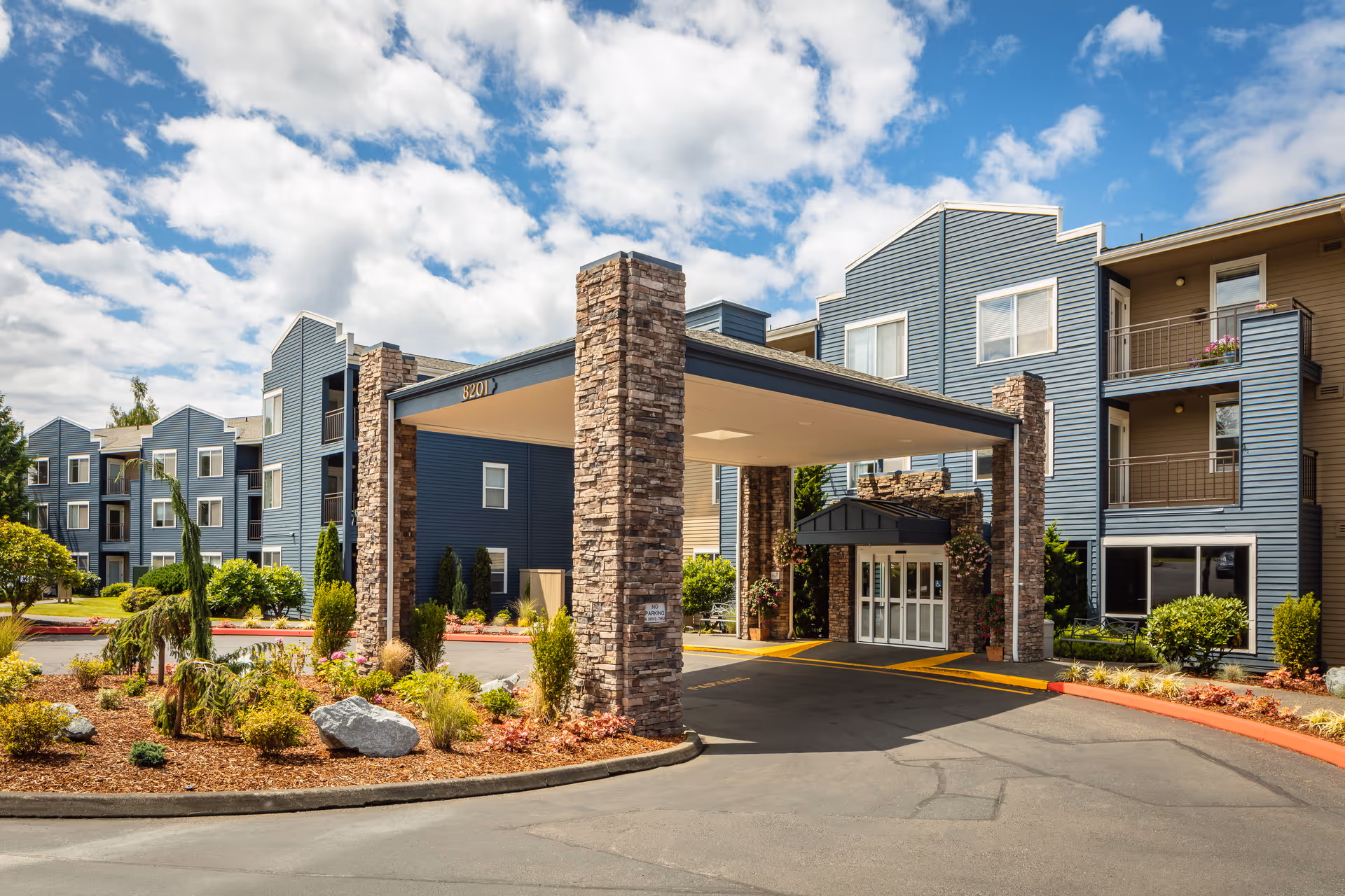 Exterior view of Cogir at The Narrows senior living facility showing a covered entrance with stone pillars, blue siding buildings, balconies, landscaped garden beds, and a partly cloudy sky.