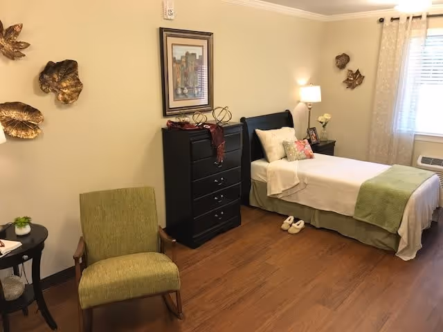 A cozy bedroom in a senior living facility with a single bed made with white and green bedding, a black chest of drawers, a green armchair, a small round side table with a plant, wall decorations including a framed picture and metallic leaf sculptures, a floor lamp, and a window with sheer curtains letting in natural light.