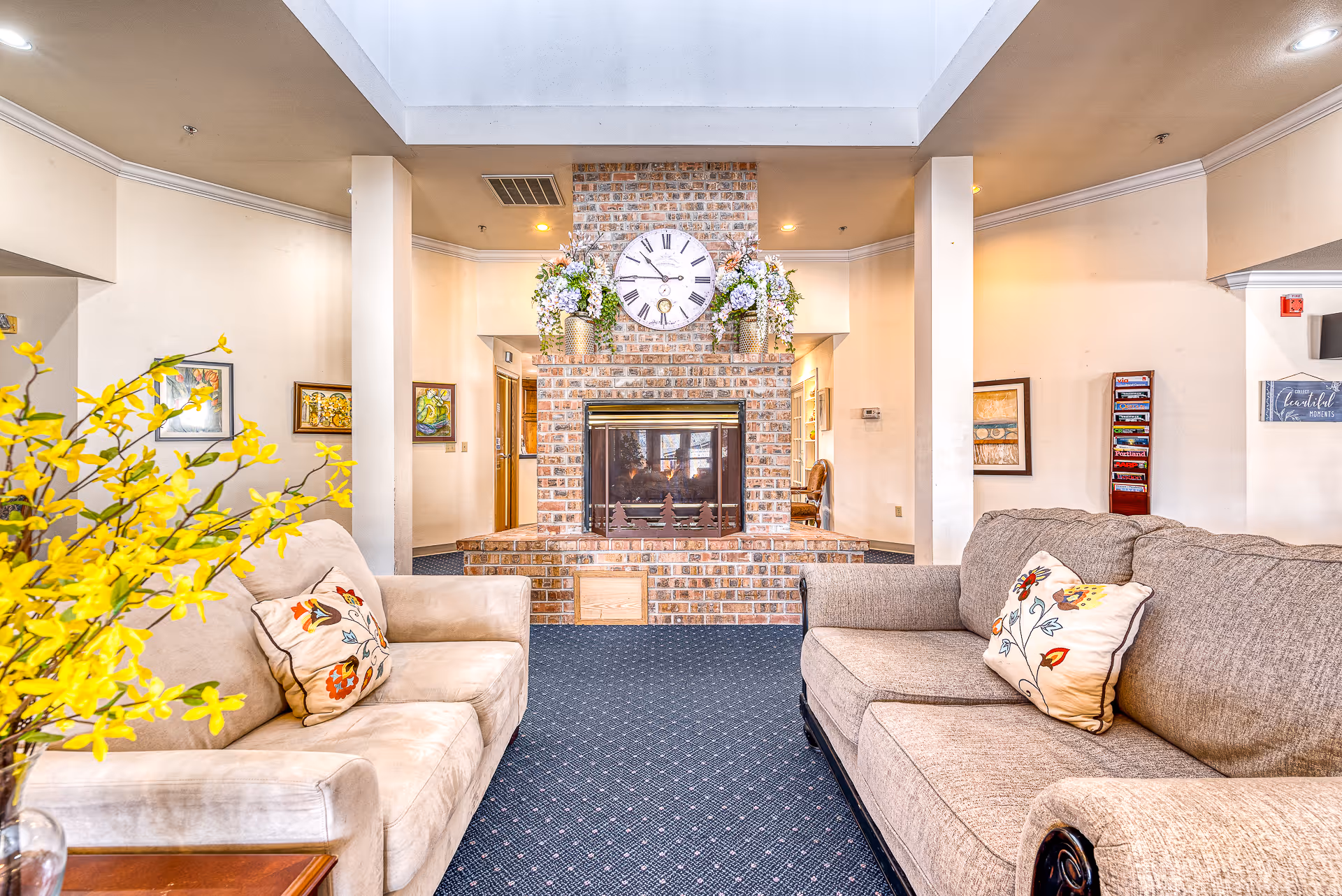 A cozy living room area in an assisted living facility featuring two beige sofas with floral patterned pillows facing a brick fireplace. Above the fireplace is a large round clock and two vases with floral arrangements. The room has a blue carpet with a subtle pattern, light-colored walls, and several framed artworks. A skylight above provides natural light.