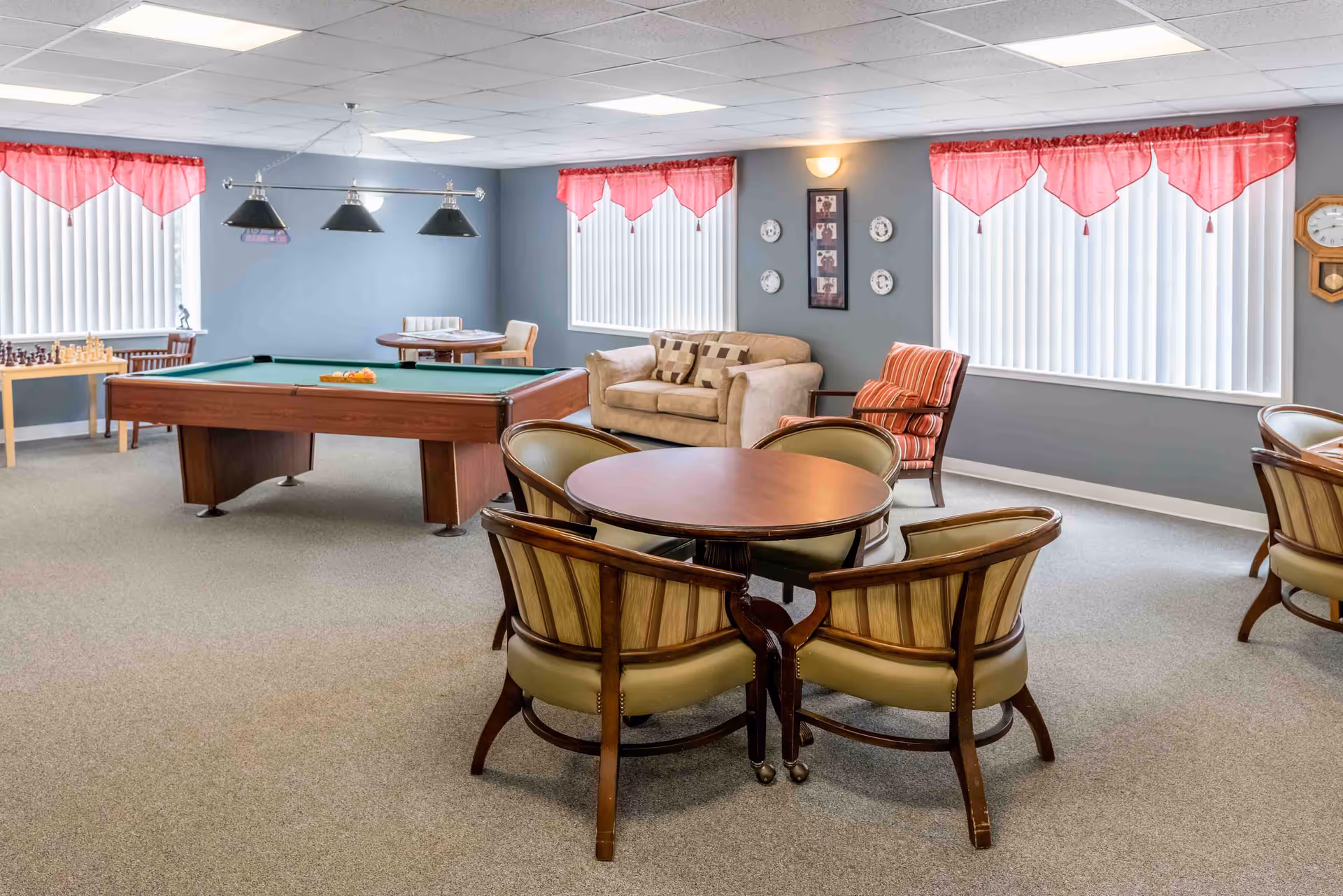 A recreational room with a pool table, a round table with four chairs, a beige loveseat, a striped armchair, and windows with vertical blinds and red valances. There is also a chess set on a small table near the window and a wall clock on the right side.