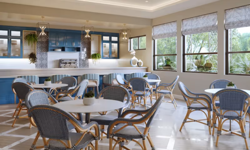 A bright dining area with round white tables and wicker chairs with blue cushions. The room features large windows with patterned blinds letting in natural light, blue cabinetry with glass doors, hanging pendant lights, and decorative plants on the counter and tables.