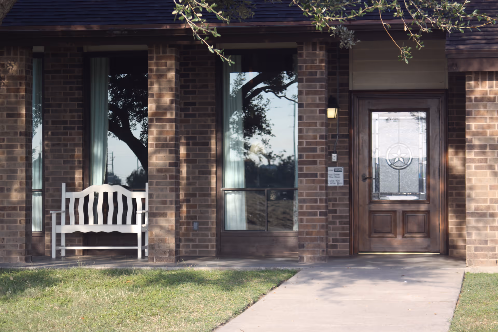 Front entrance of a brick nursing facility with a wooden door, large windows, and a white bench on the covered porch.