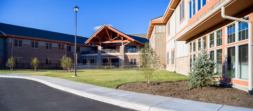 Exterior view of The Foley Center at Chestnut Ridge showing a modern two-story building with large windows, brick and stone facade, a covered entrance with wooden beams, a well-maintained lawn, small trees, and a sidewalk under a clear blue sky.