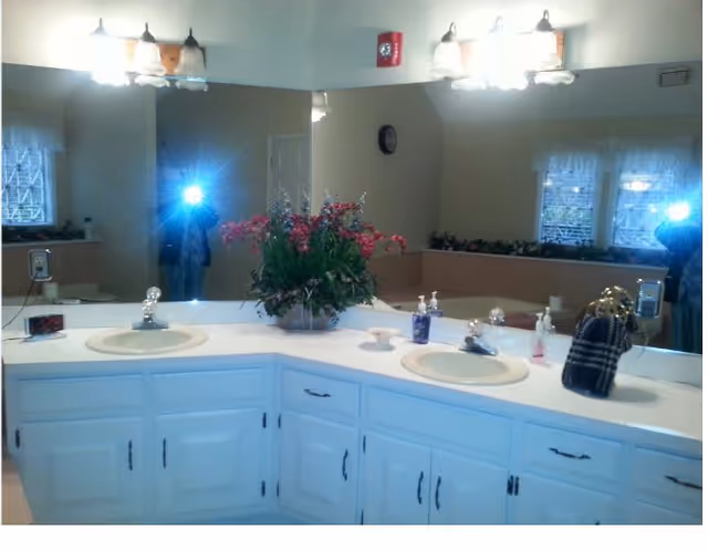 A bathroom vanity with two sinks, each with a faucet and soap dispensers. There is a large mirror behind the sinks reflecting the room, including a clock on the wall and windows with decorative curtains. A floral arrangement is placed in the center of the countertop. The lighting fixtures above the mirror have multiple bulbs with frosted glass shades.