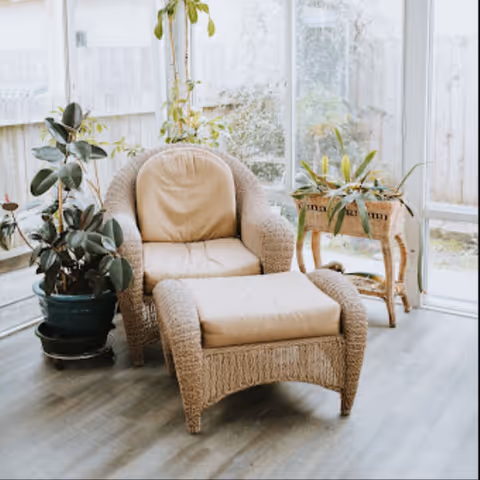 Sunlit glass-walled living room with a wicker chair, ottoman, and potted plants.