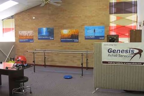 Interior rehabilitation room with wall posters, a rail, desk, and Genesis Rehab Services sign.